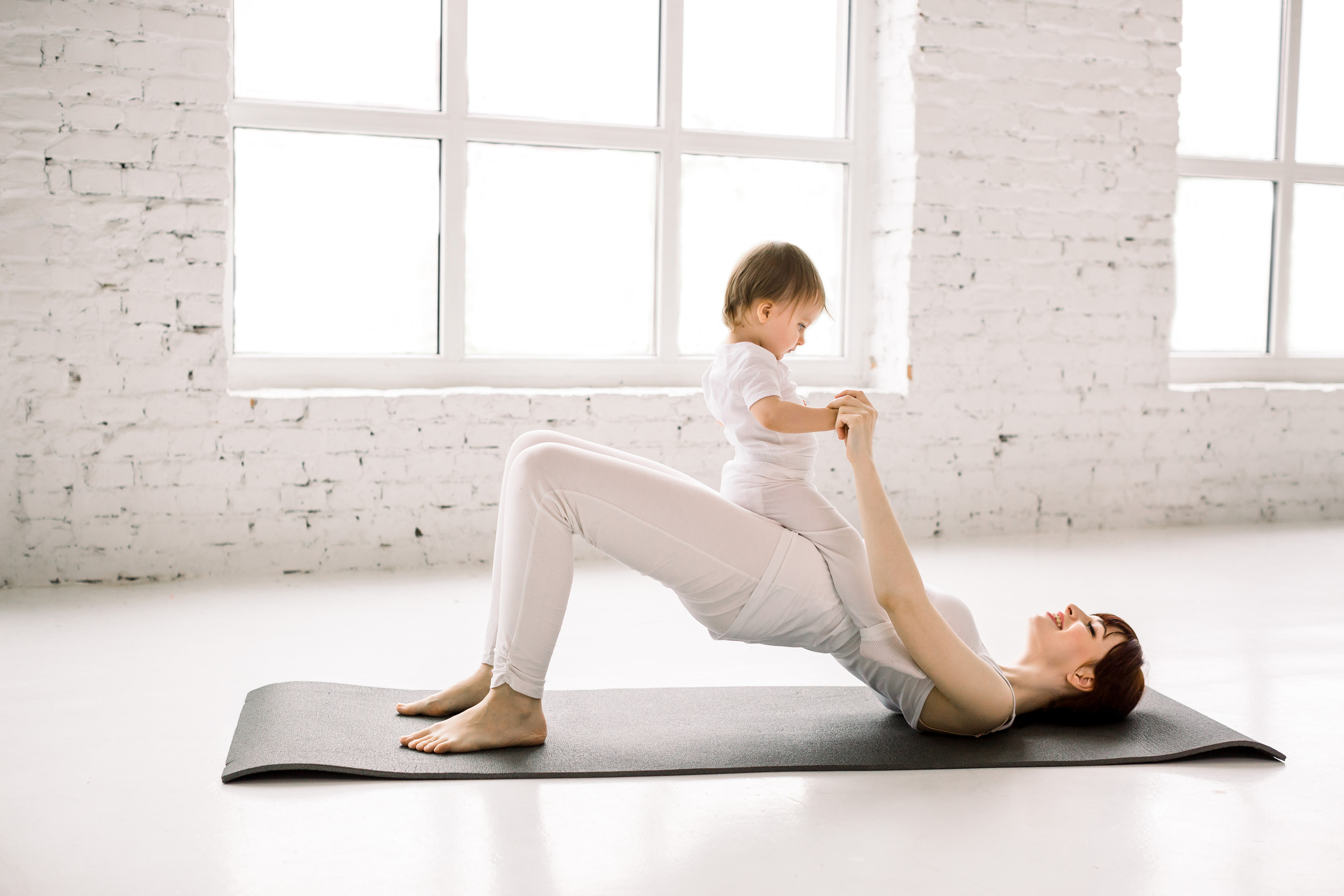 Young happy mother working out, doing butt bridge exercise, wearing white sportswear, little baby girl on her tummy, fitness, postnatal yoga. Healthy lifestyle concept